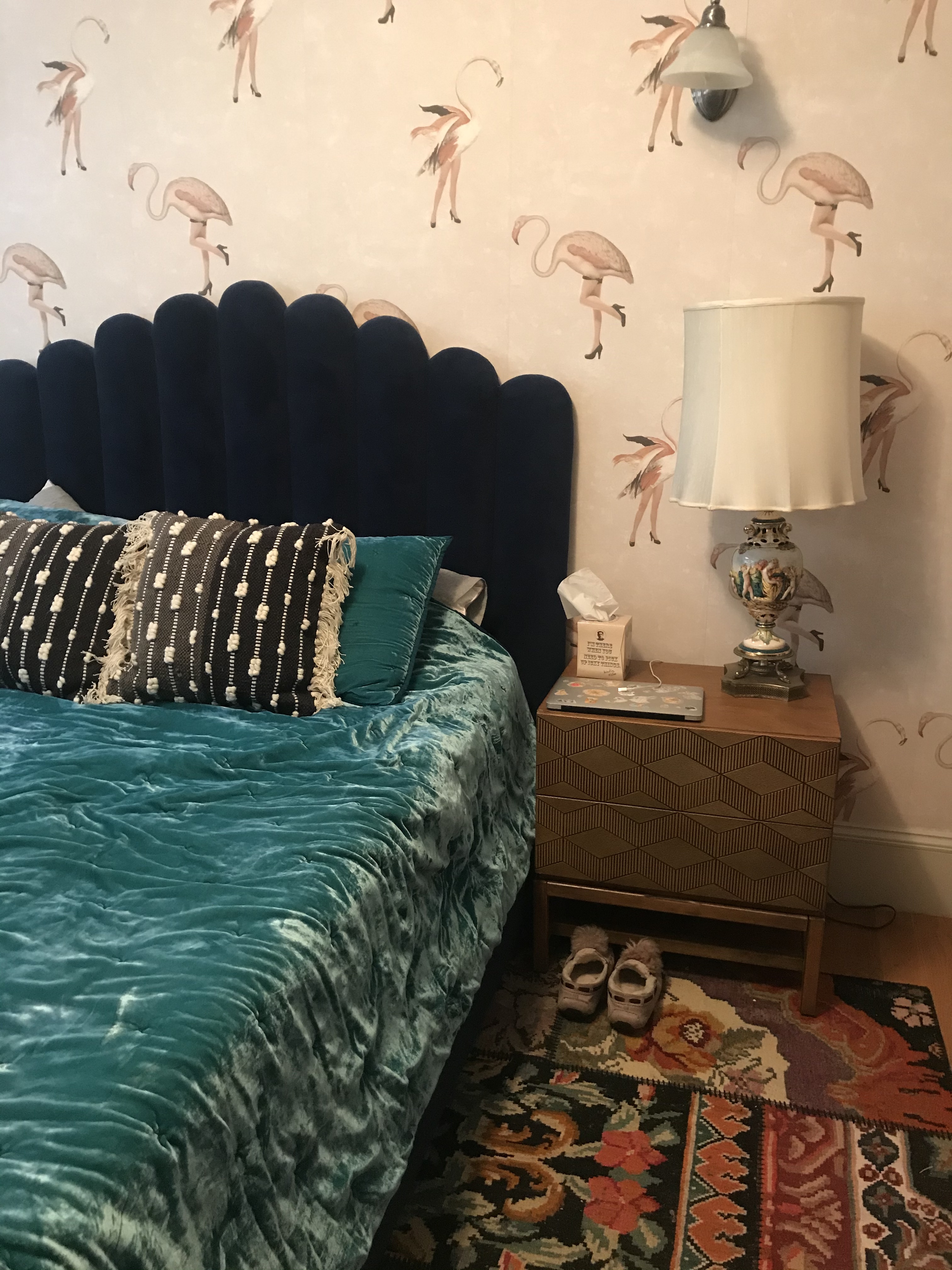 Bedroom with pale pink wallpaper covered in flamingos with human legs wearing black high heels, a dark blue headboard, a deep turquoise velvet bedspread, colorful rug, and nightstand with a lamp