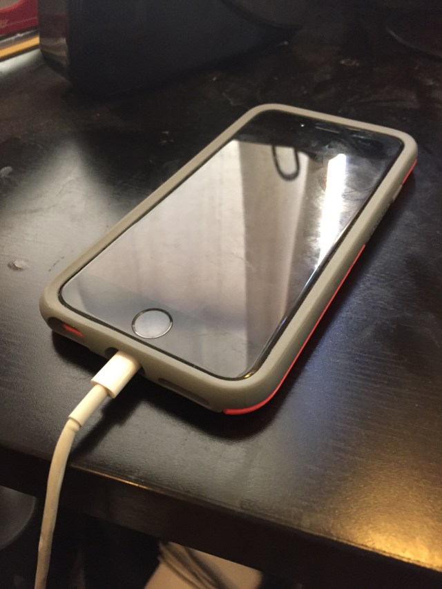 A black iPhone 6s with a pink case sits plugged in on a black desk.