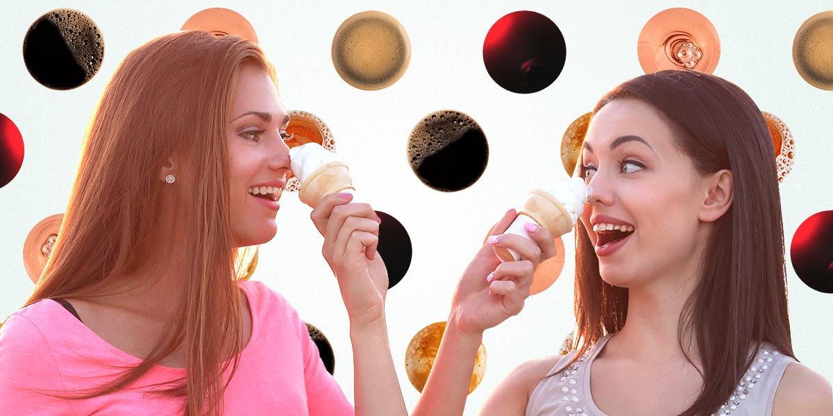 Two women pushing ice-cream cones on each other's noses and laughing, background is a pattern of various beverages