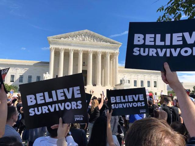 Protestors gathered outside the U.S. Supreme Court, images of three black and white signs that say "Believe Survivors" in the foreground with the Supreme Court in the background against a blue sky.