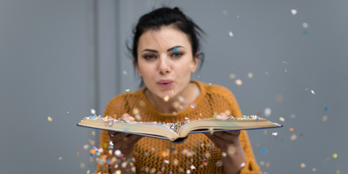 woman in a mustard sweater blowing confetti off an open book at the camera