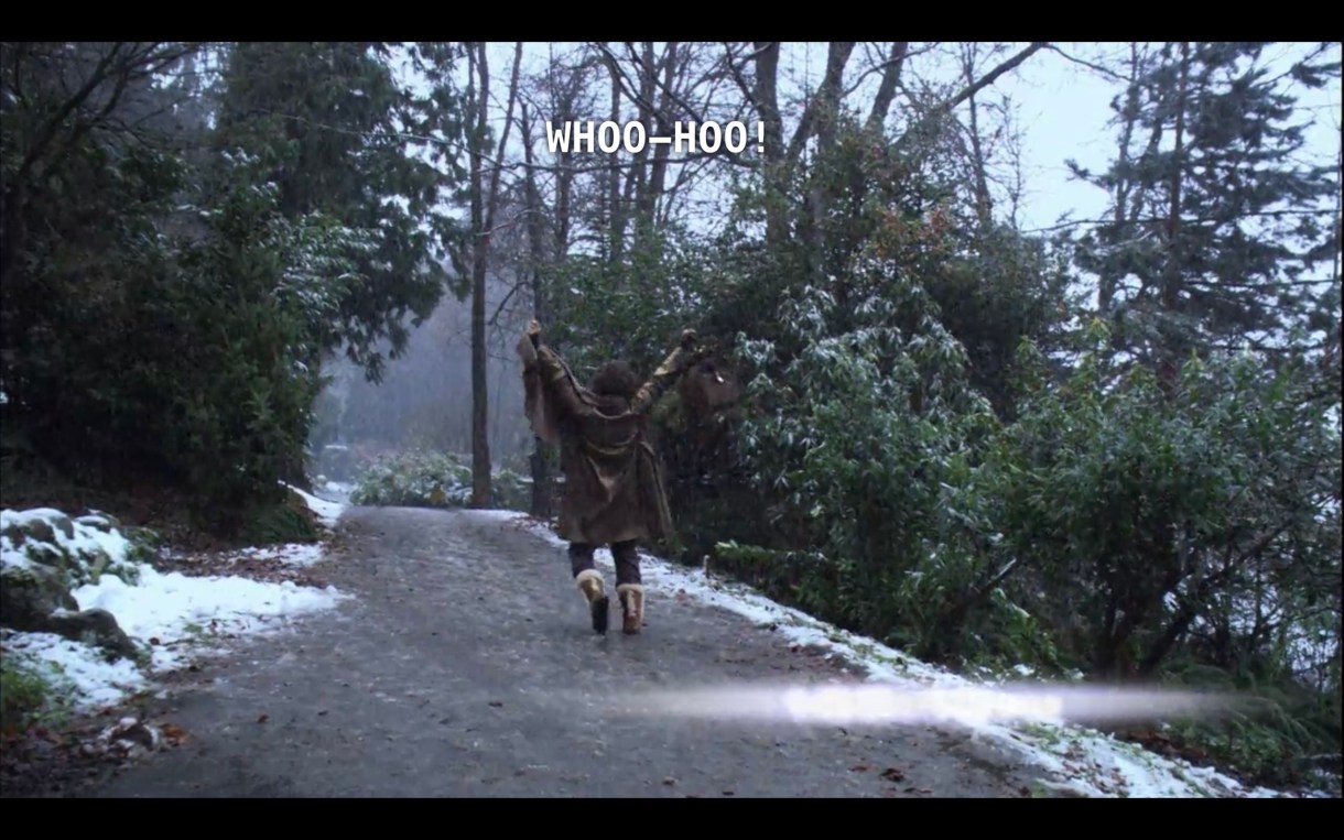 Bette, with her back to the camera, is walking down a dirt path in the woods, with a light dusting of snow. She raises both arms in the air and yells, "Whoo-hoo!" as she leaves her silent retreat.