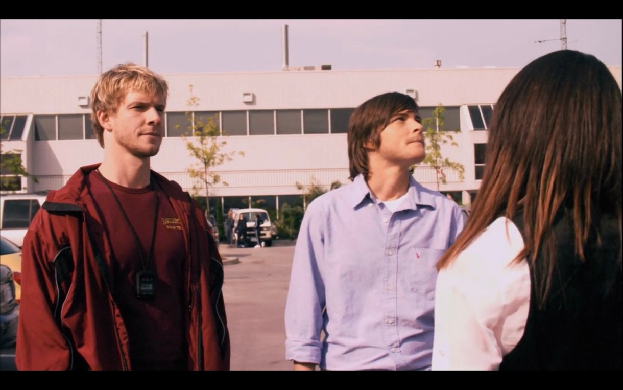 Tim (wearing a red t-shirt under a red track jacket) stands next to Max (wearing a blue button-up) and opposite of Jenny (whose back is to the camera) in a parking lot. Tim is looking blankly at Jenny, and Max is looking embarrassed and off into the distance.