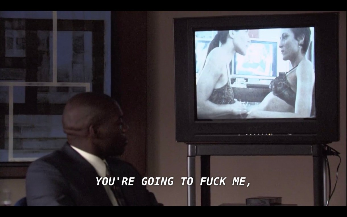 A bald man in a full suit looks back over his shoulder at an old chunky TV on a rolly cart. On the TV is a blurry video of Helena and Dylan in their bras. Through the TV Helena says, "You're going to fuck me."