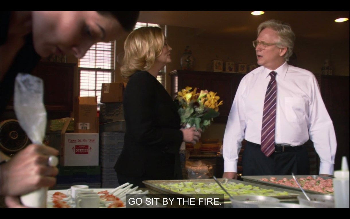 Phyllis (in a black blazer) stands in a commercial kitchen, telling an old white guy in a white shirt and red tie, "Go sit by the fire."