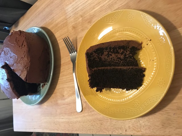 A homemade double chocolate layer cake. In the foreground is a slice of the cake on a yellow ceramic plate with a silver fork. In the background is the full cake (with the slice missing) on a light mint green plate. Both plates sit on a long wooden table.