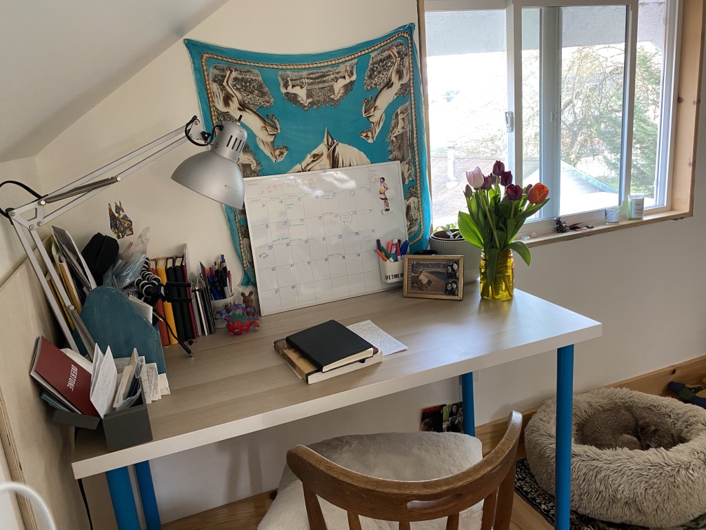 Camila's well-organized desk featuring a vase of flowers, a large calendar, multiple notebooks, and a dog bed beside it