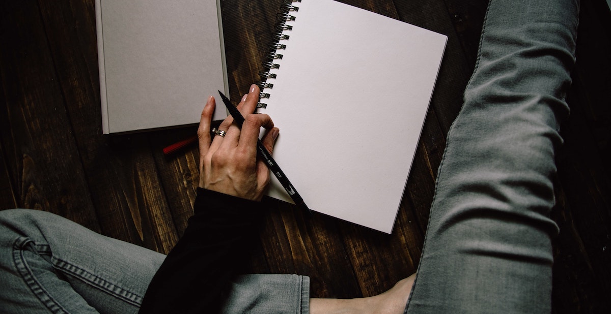 a person sits with one leg crossed and the other outstretched and a pad of paper and a notebook on the floor in front of them, pen poised as if about to write or sketch