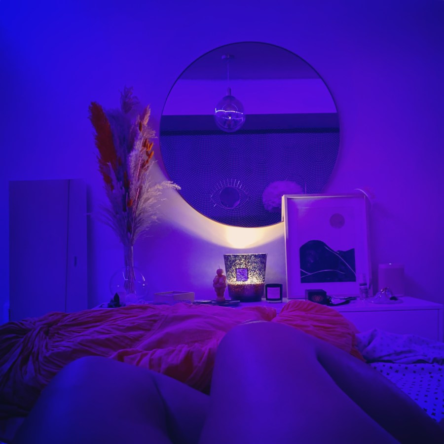 Deep blue and purple light fill a room with a round mirror on the wall, feathers and prints on either side, with legs on a bed in the foreground