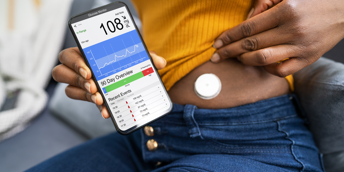 A Black person wearing a yellow top and jeans lifts up their shirt to reveal a Continuous Glucose Monitor in their abdomen. They hold up their phone, which shows a glucose monitoring app.
