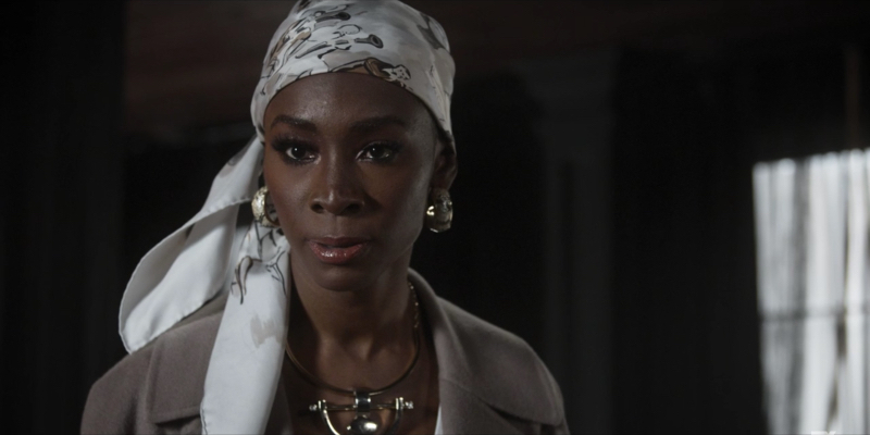Angelica Ross looks serious with a white headwrap, chunky white earrings, and a tan jacket