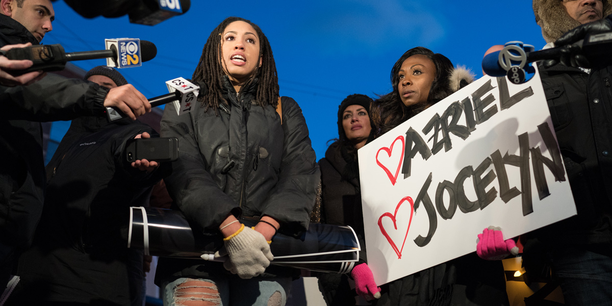A group of protestors stand outside R. Kelly's hearing, a black woman speaks into a microphone as another black woman holds a sign that says "Ariel and Jocelyn, two of R Kelly's victims, with hearts around them"