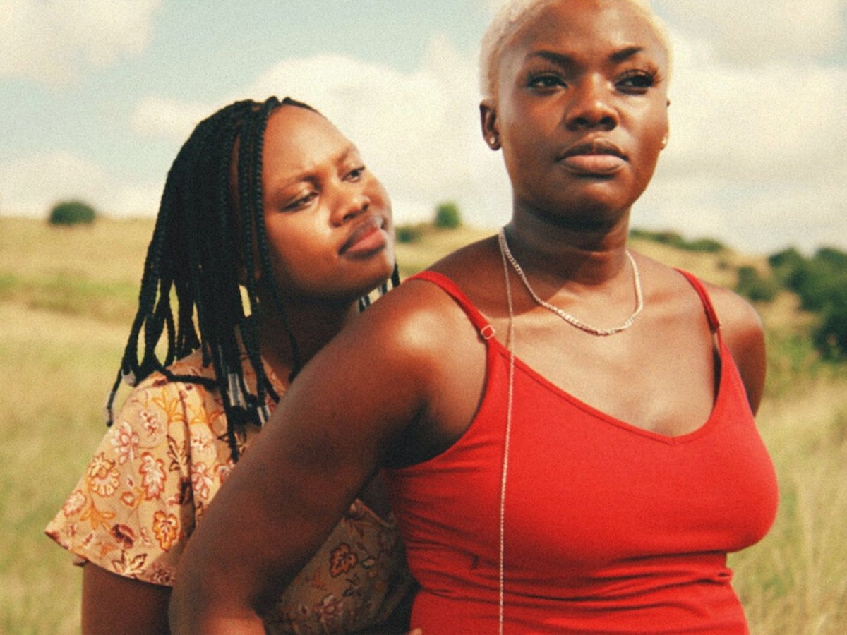 two Black women in a gorgeous field, one in a red tank top with a shaved head and the other with locks standing behind her in a yellow paisley shirt
