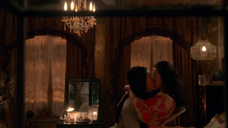 Alice and Sumi share a kiss in Sumi's loft. Sumi is in her robe and Alice is in a grey suit. There's a huge chandelier to their left.