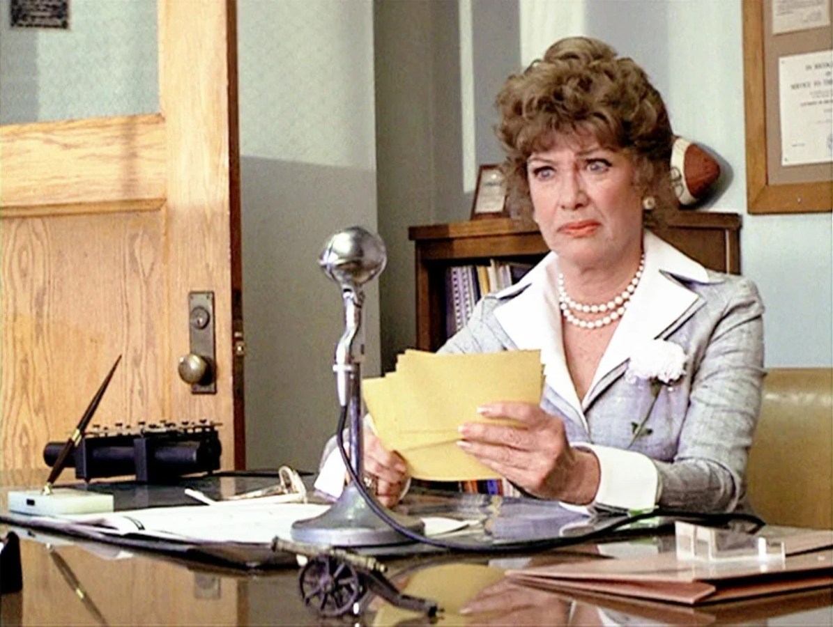 Principal McGee at her desk at Rydell High in front of a microphone, shuffling notecards