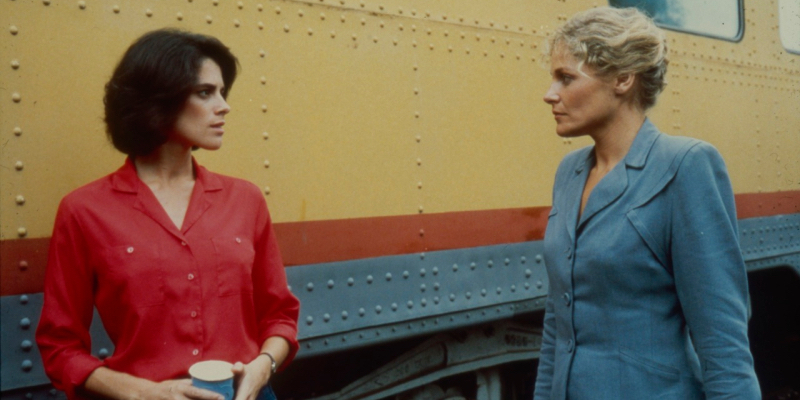 A middle aged woman in a blue suit looks at a younger woman in a red top by a train.