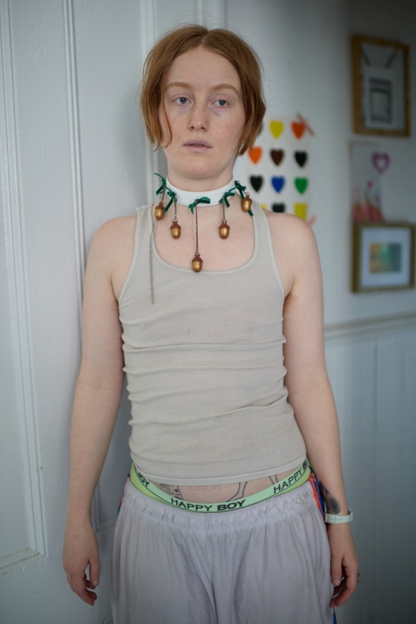 Bobbi stands in a doorway in a tank top with acorn bells around their neck