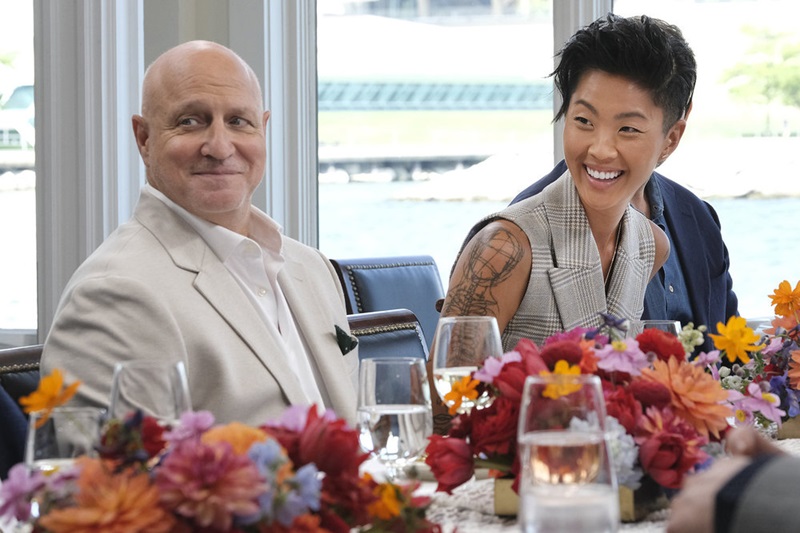 Top Chef judges Tom Colicchio and Kristen Kish react to commentary from another (off-screen) dinner guest. Kristen is smiling brightly, wearing no sleeves and her tattoo on her right arm is fully visible.