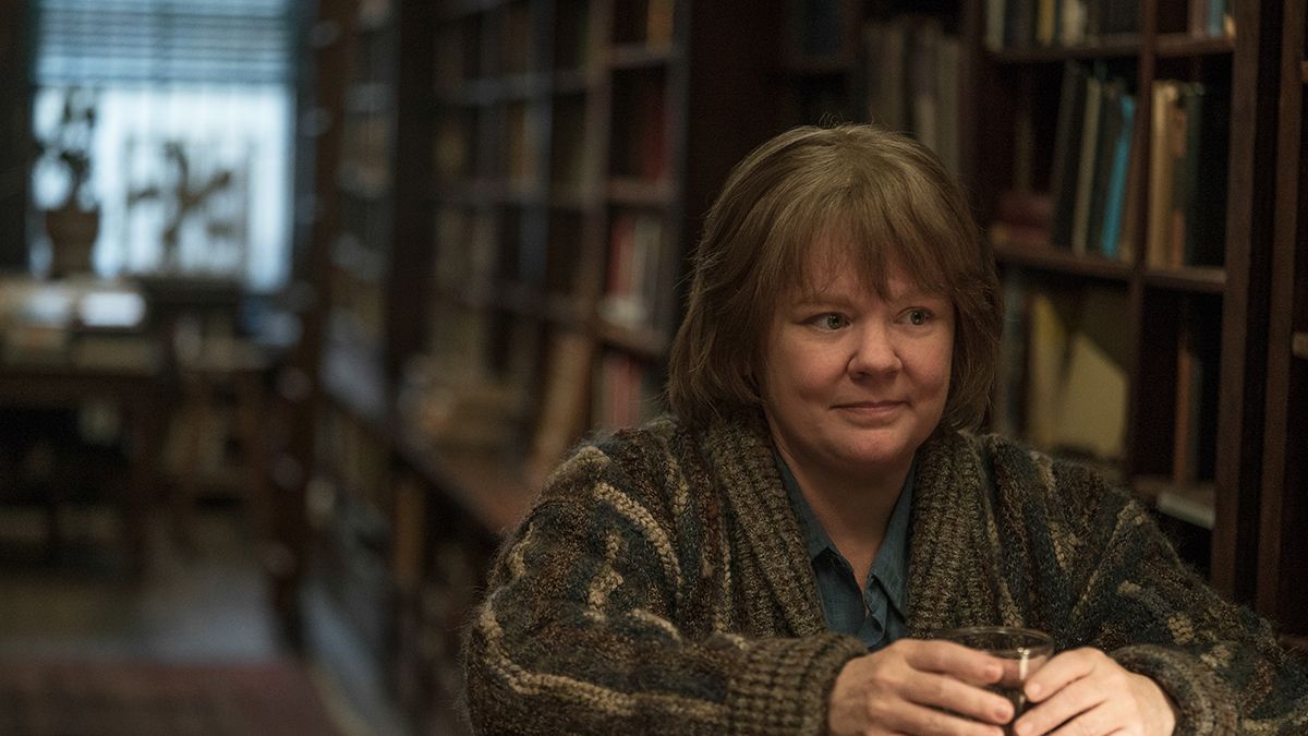 Melissa McCarthy stands in a bookstore with a glass of whiskey. 