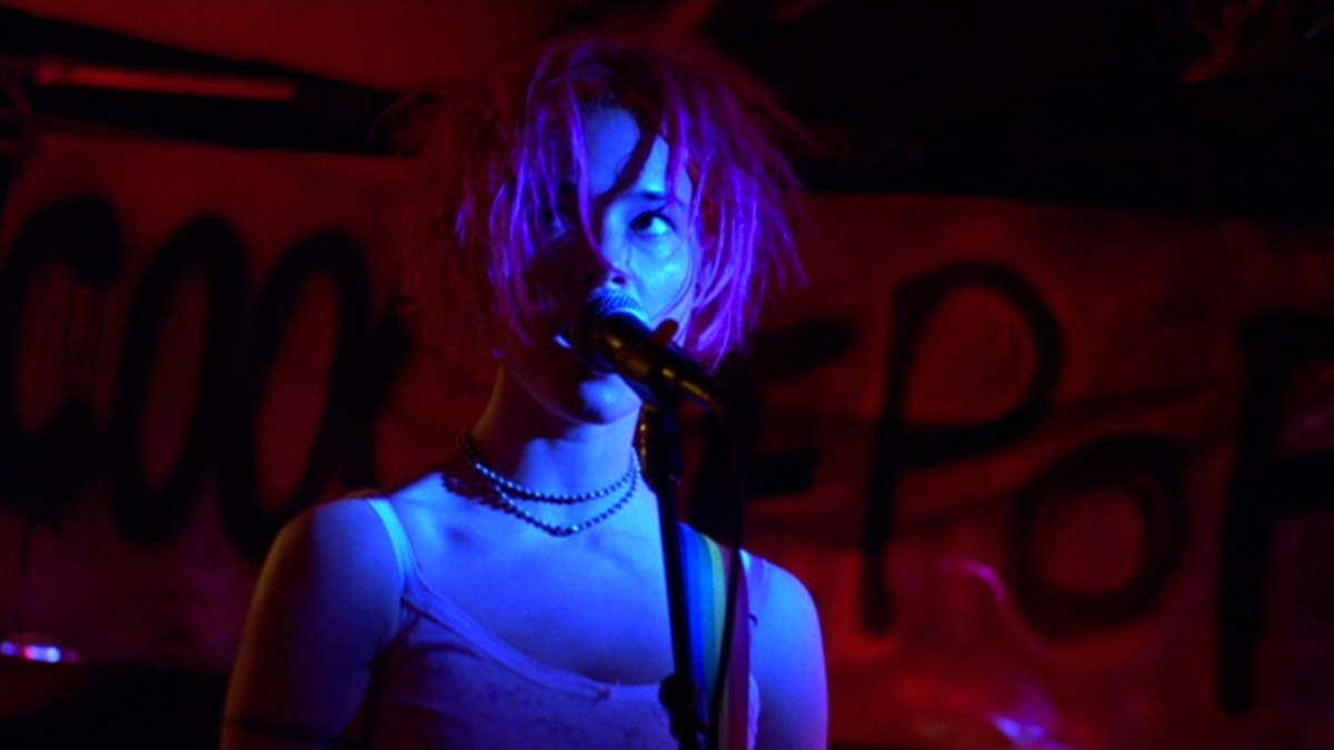 Leisha Hailey with pink hair stands in front of a microphone on stage