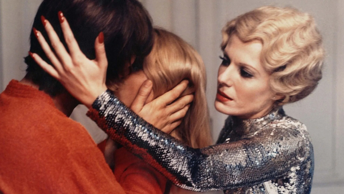 Delphine Seyrig with blonde hair holds the back of a man's hair as he kisses a woman