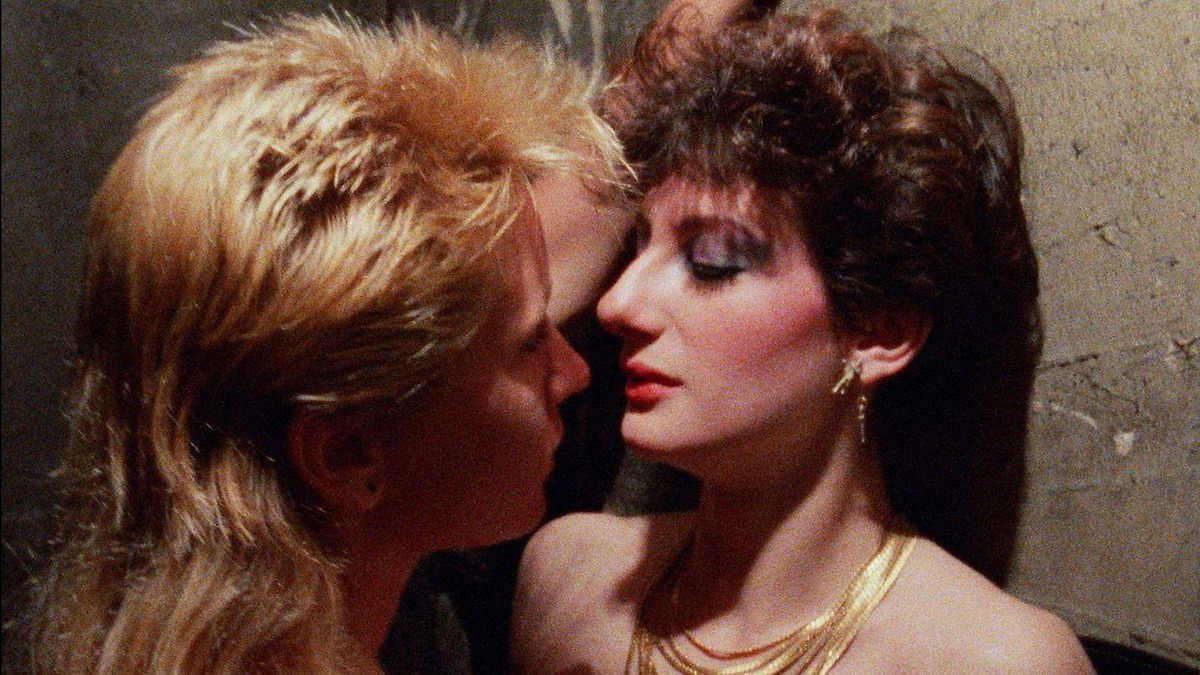 In a screenshot from Kamikaze Hearts, two women with mullets (one blonde, one brunette) are pressed up together against a wall. The blonde mullet has her arms grazing the shoulder and face of the brunette. It's the 1980s, with 1980s style bright make up included.