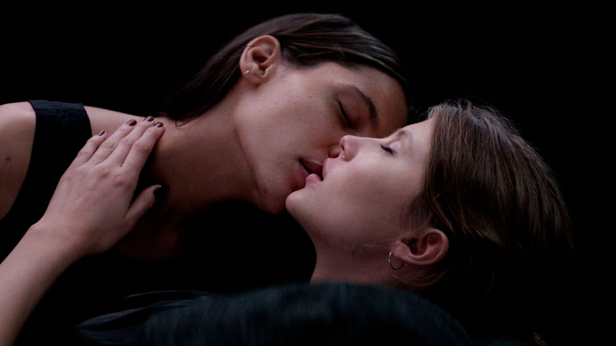 Two young women kiss against a black background.