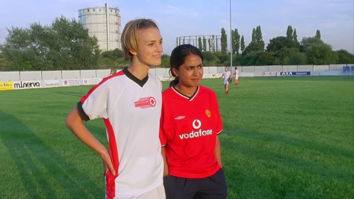 Keira Knightley and Parminder Nagra stand next to each other in soccer jerseys. 