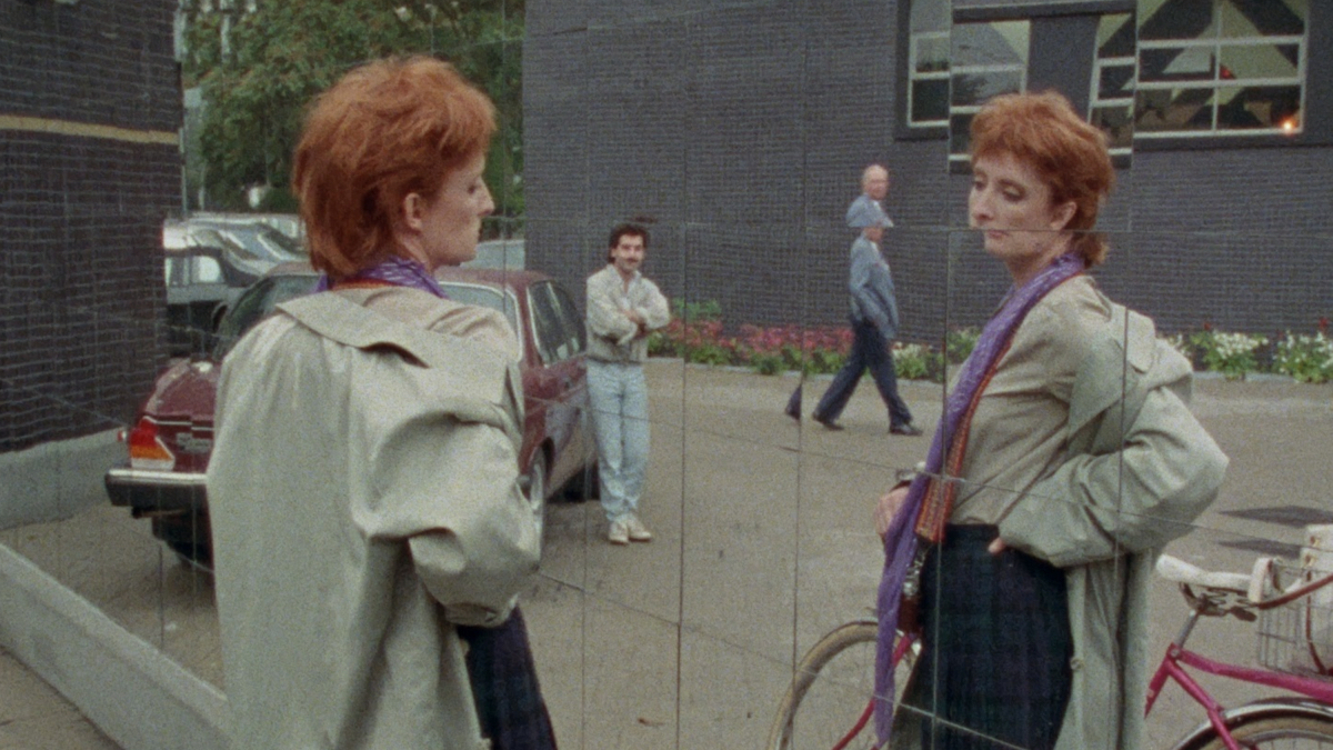 A woman with short red hair looks at her refracted reflection in a tiled mirror wall.