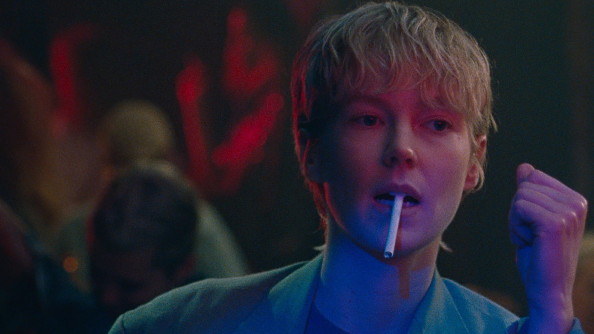 A close up of a blonde woman with short hair sits at a bar with a cigarette dangling from her mouth.