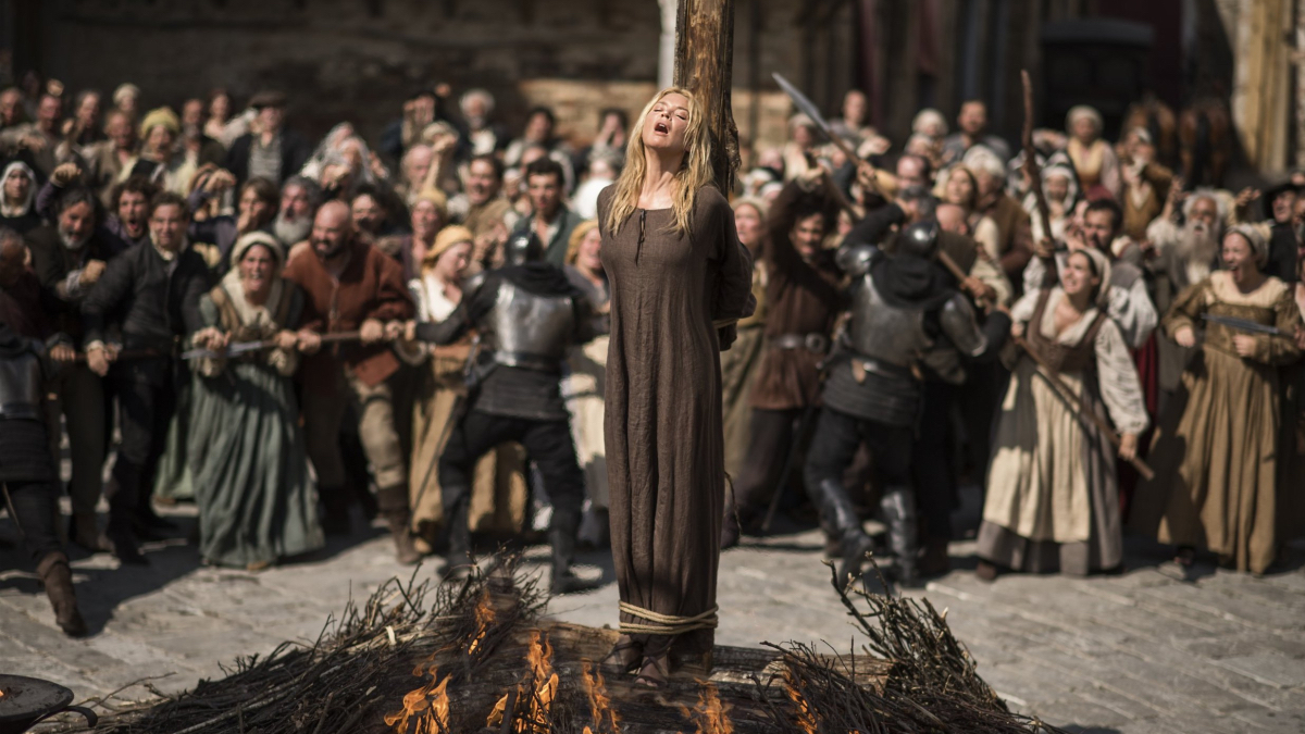 Virginie Efirie looks up while tied to a wooden stake being burned while a crowd gathers behind her.