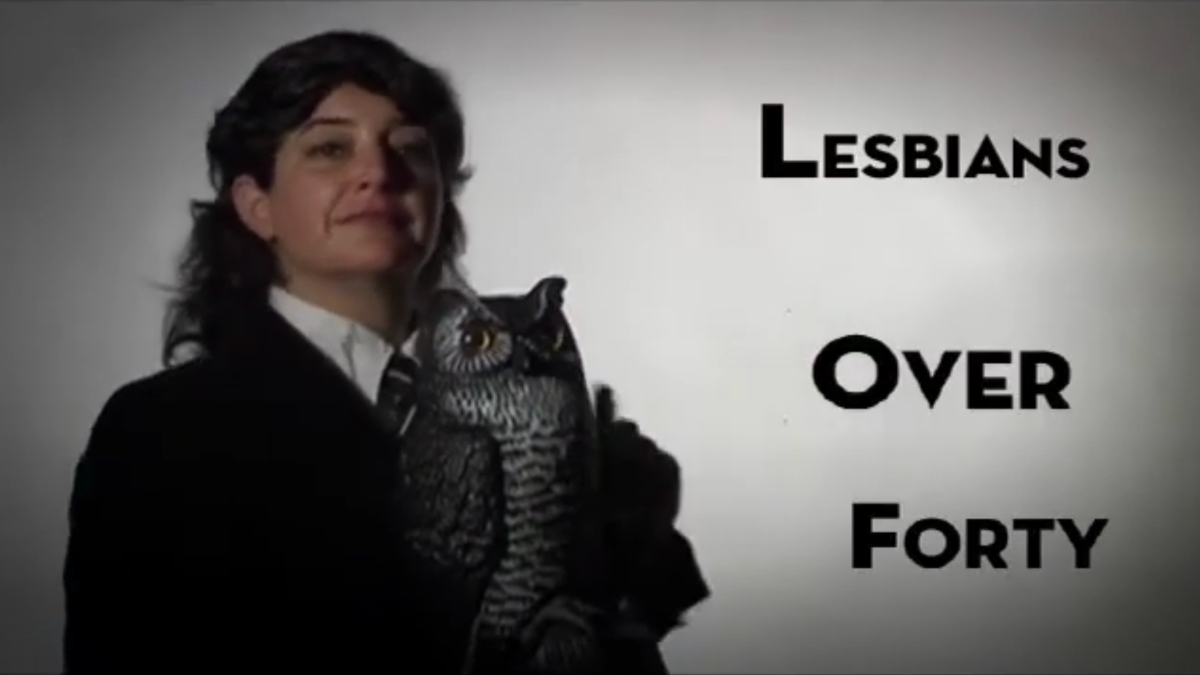 Close up of Anna Margarita Albelo holding a fake owl next to text that says Lesbians Over Forty