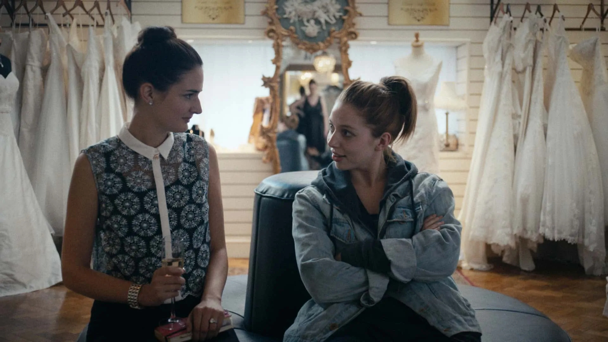 A woman in a jean jacket sits with her arms crossed and looks at her friend in a wedding dress shop