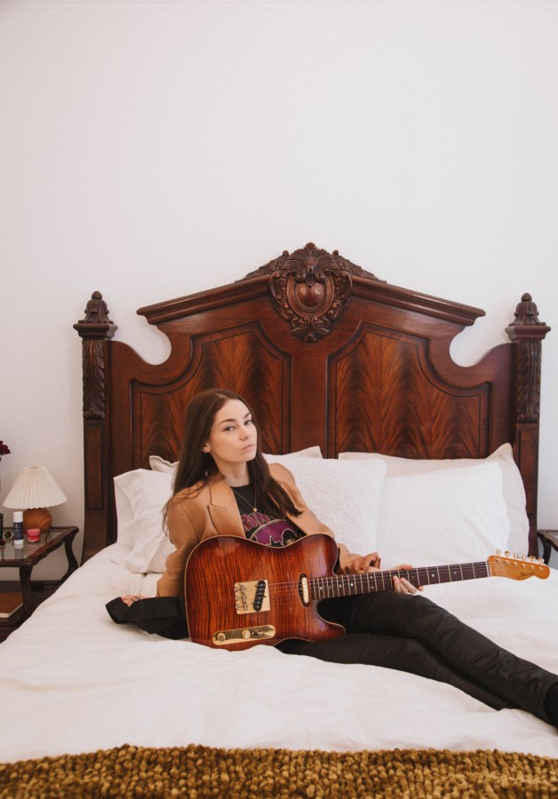 She lies on a bed with a wooden backboard holding her guitar.