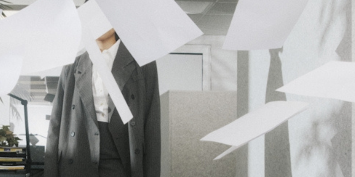A woman in an oversized suit stands in an office space. Blank sheets of paper flutter around covering her face.