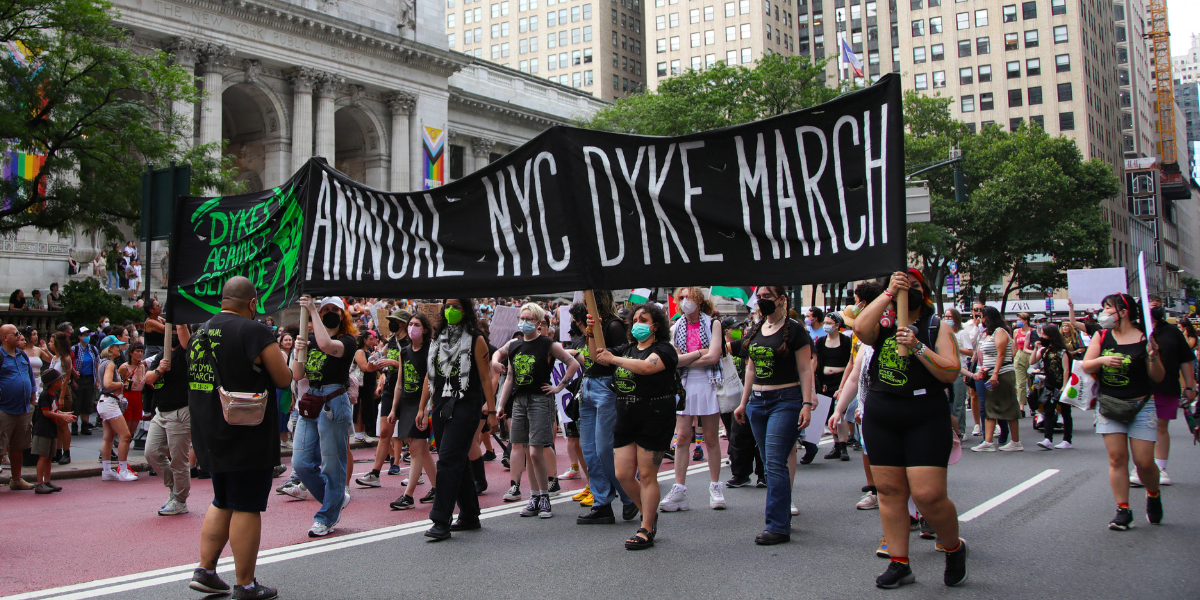 A group of queer people in black and lime green t-shirts and black masks hold up a big sign that says Dykes Against Genocide Annual NYC Dyke March