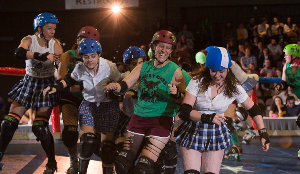 A group of queer smile as they bump into each other on a roller derby rink.