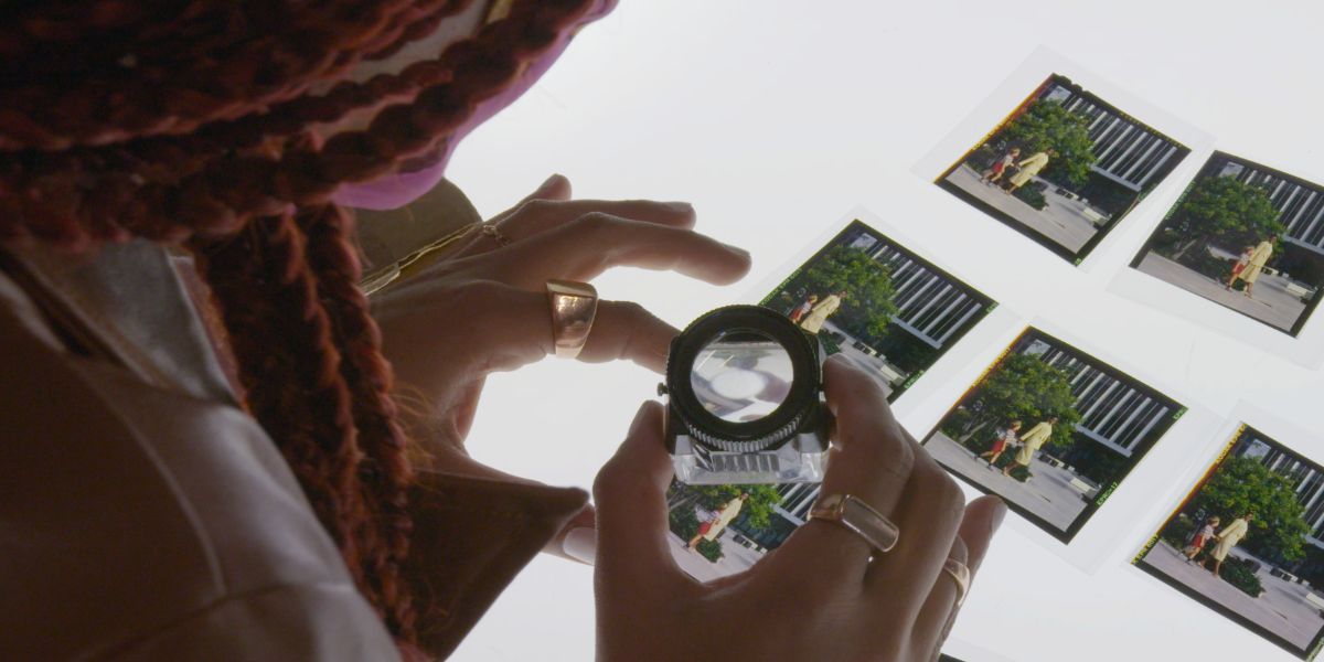 A Black femme leans over an old photograph with a microscope in Seeking Mavis Beacon