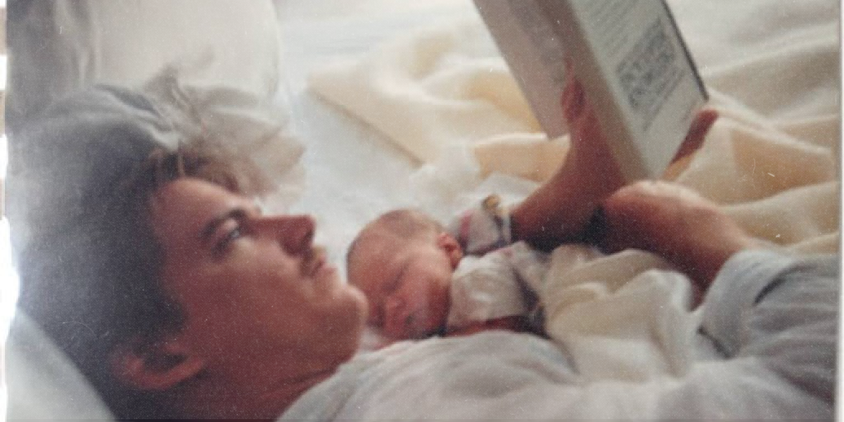 A photo from the 90s of a man with a mustache reading a book with a baby on his chest.