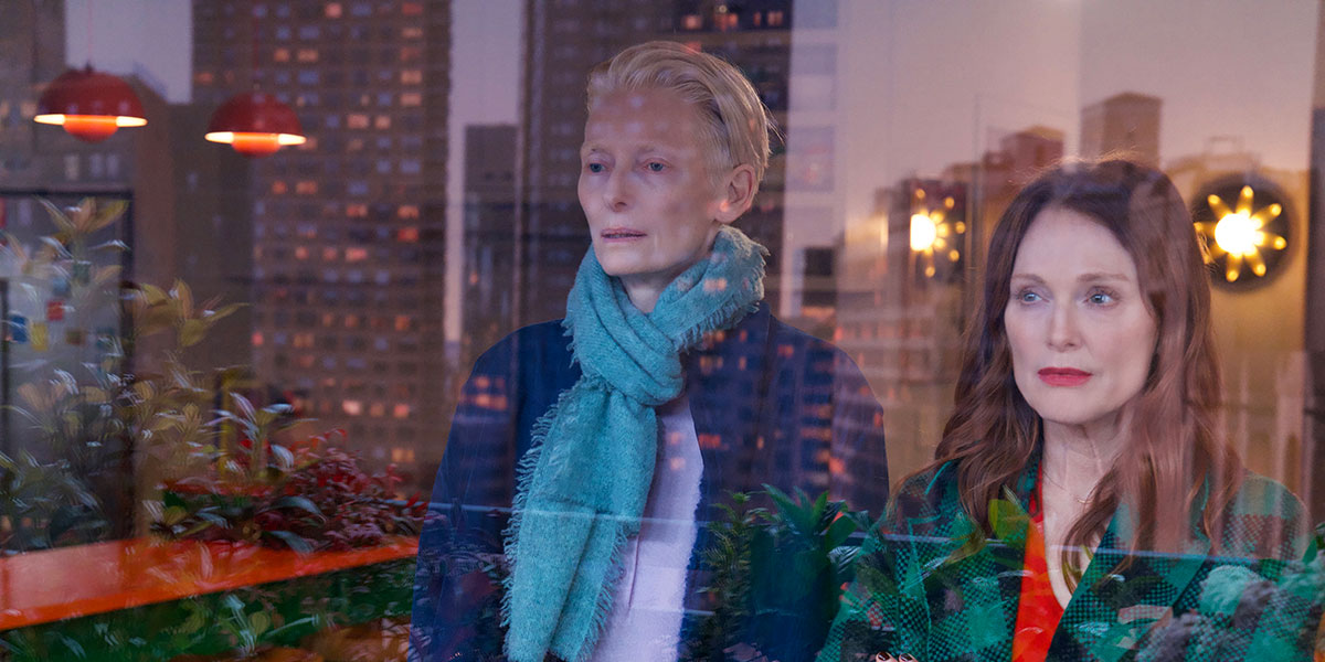 Tilda Swinton and Julianne Moore stand side by side looking out a window with the cityscape reflected back at them in The Room Next Door.