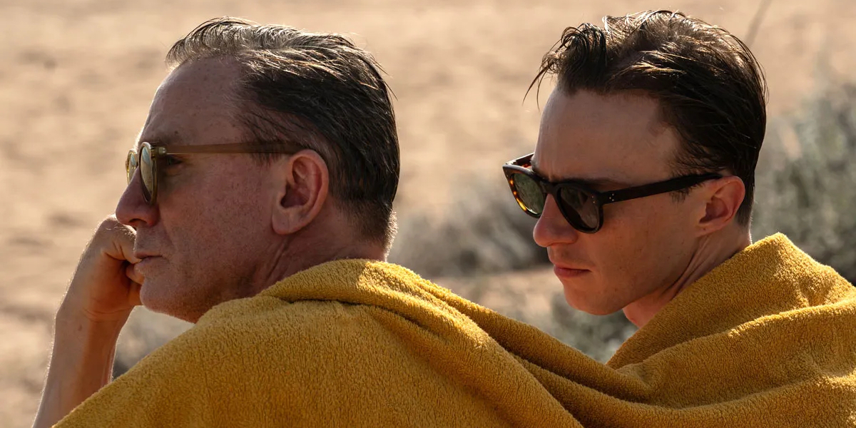 Daniel Craig and Drew Starkey sit on the beach with one yellow towel wrapped around them both wearing dark sunglasses.