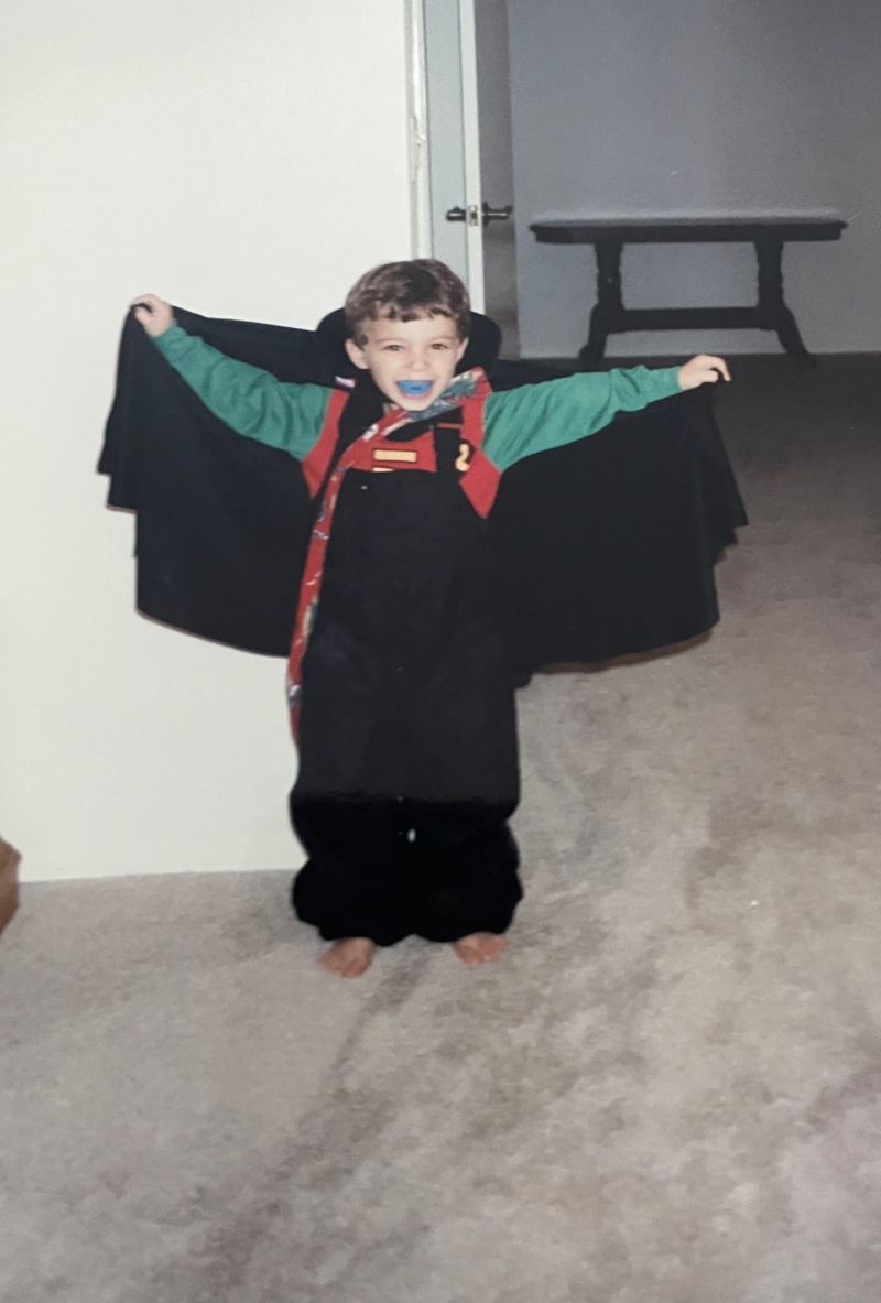 Drew, age 4, lifts her Dracula cape she wears over a Robin costume with blue plastic fangs.