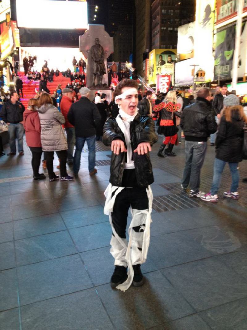 Drew, age 18, stands in Times Square with her arms outstretched wearing a leather jacket dressed as a mummy.