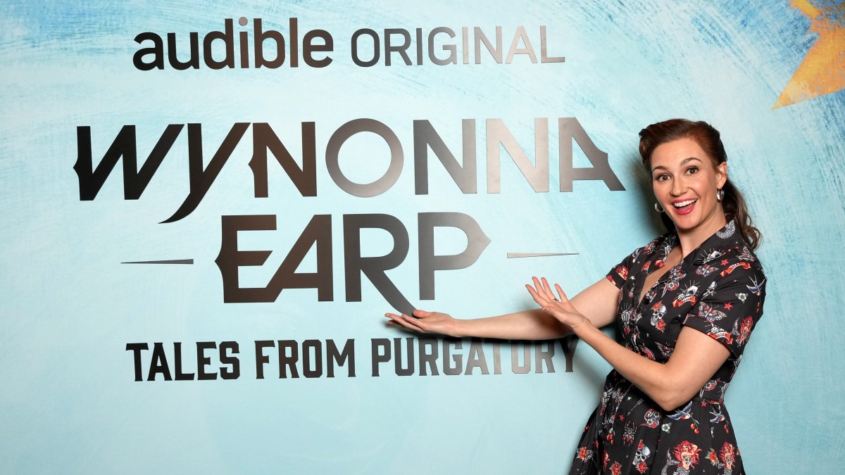 Kat Barrell next to a sign that says "Audible Original Wynonna Earp Tales from Purgatory"