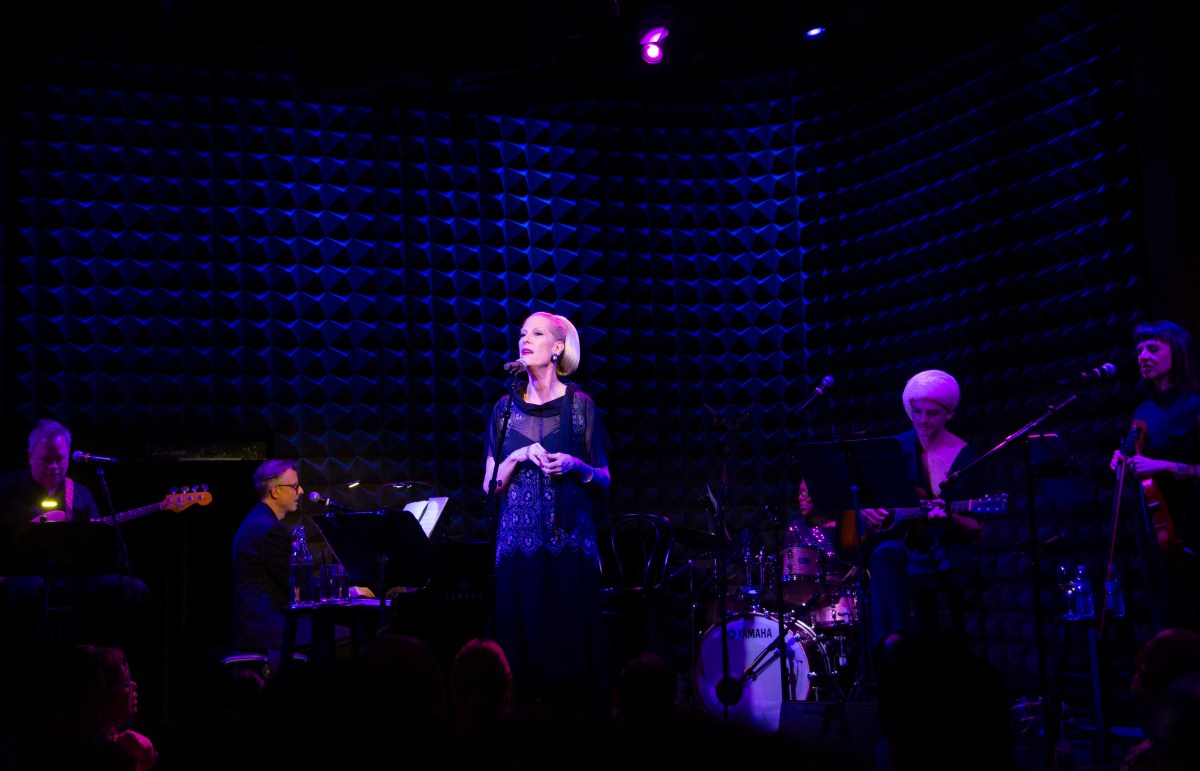 A wide shot of Justin Vivian Bond singing on stage with their band behind them.