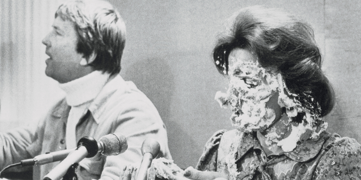 A black and white image of Anita Bryant with pie on her face as a man reacts in the background.