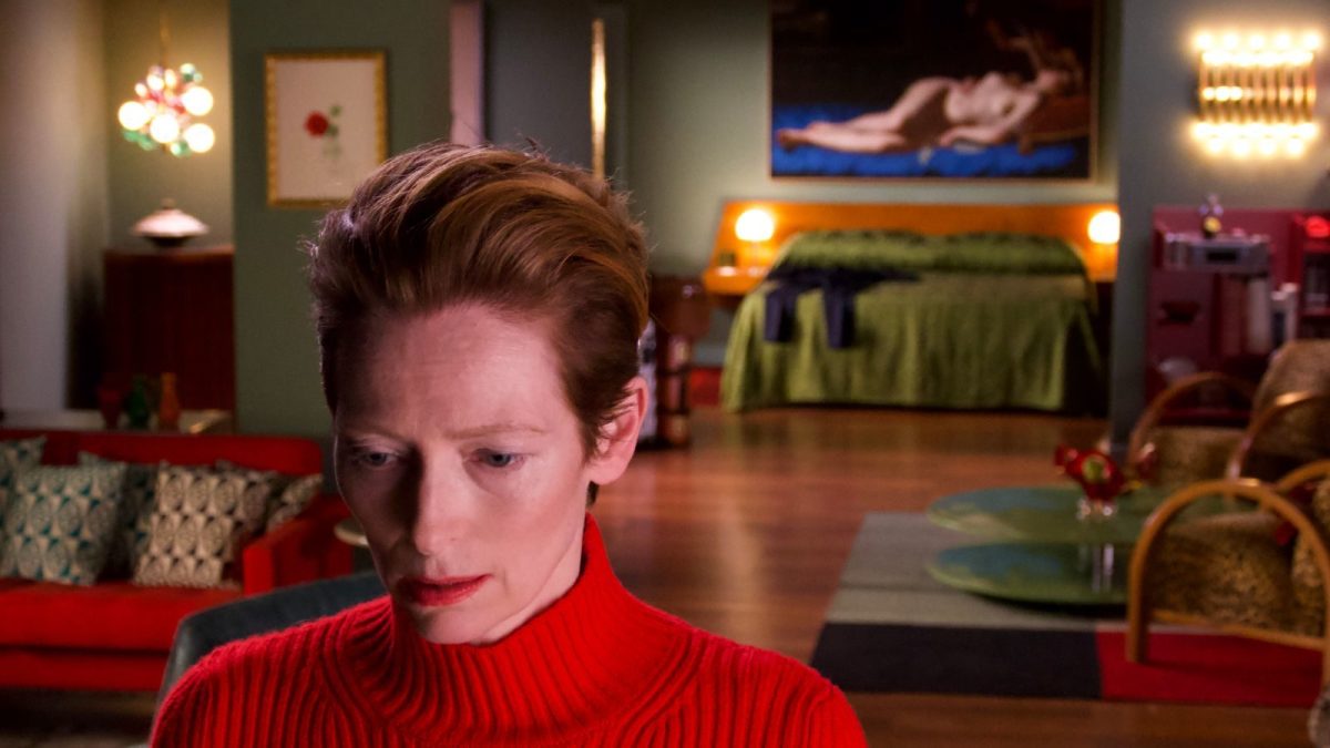 A close up of Tilda Swinton in a red sweater in the bottom left corner with her fancy apartment behind her.