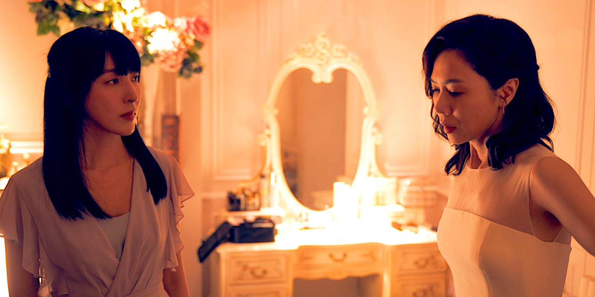 Born for the Spotlight: a woman in a pale pink top looks at a woman in a white dress in warm lighting with a vanity behind them.