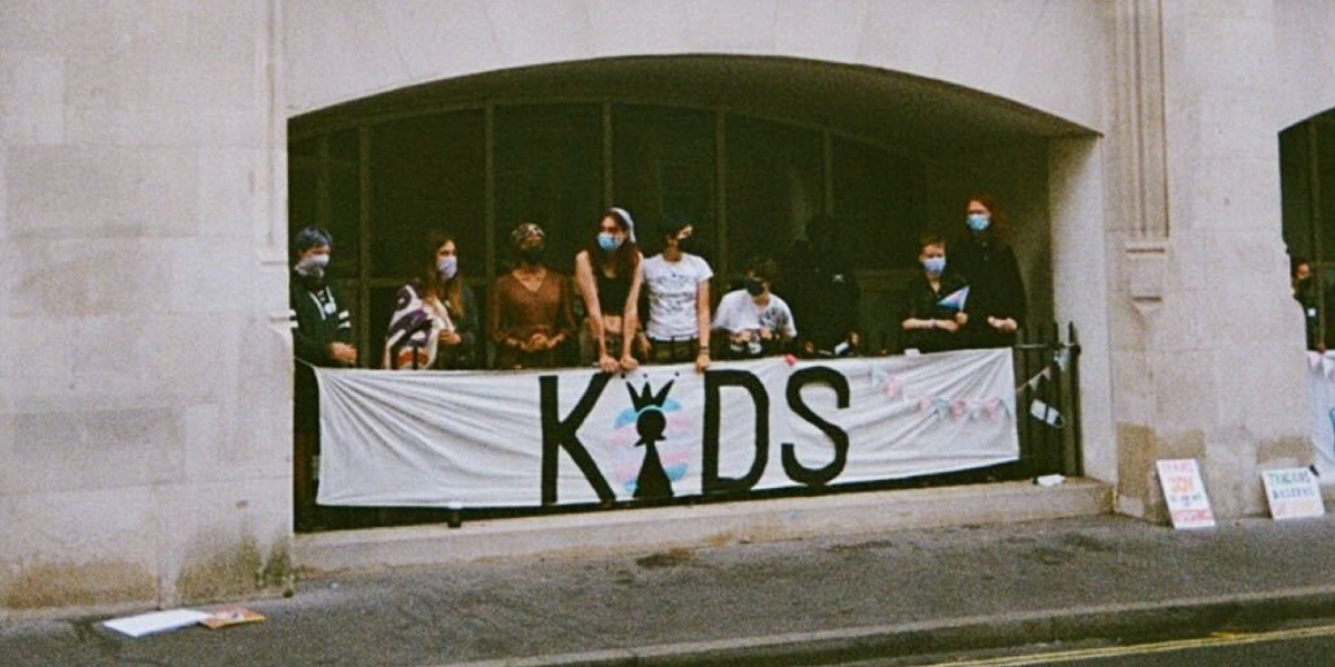 A group of trans kids wearing masks stand in an archway behind a sign that says KIDS with the i a chess piece surrounded by a trans flag.