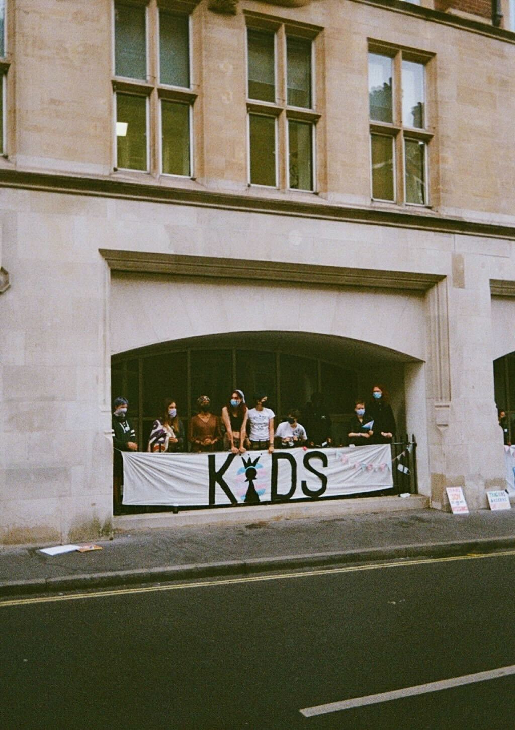 A group of trans kids wearing masks stand in an archway behind a sign that says KIDS with the i a chess piece surrounded by a trans flag.